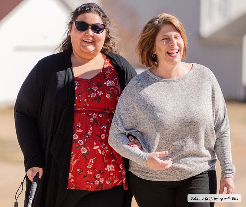 Frame-9457 A daughter and mother are walking outdoors arm-in-arm. The daughter, wearing a black cardigan and sunglasses, is holding a cane, while the mother is in a gray sweater and smiling.
