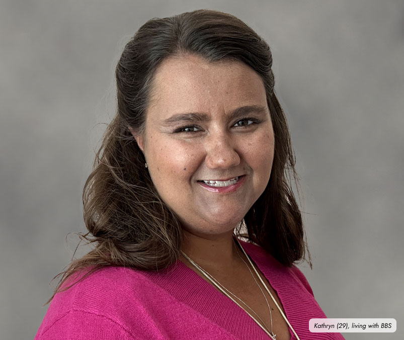 Frame-9457 A young woman, with brown hair wearing a pink top, smiles at the camera against a gray background. A small label at the bottom right of the image reads: "Kathryn (29), living with BBS and managing it with IMCIVREE.”