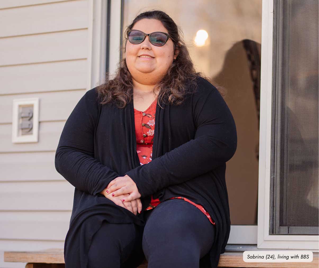 Frame-9457 A girl wearing sunglasses, and a black cardigan sits on a wooden bench near a doorway of her house. Her hands are clasped together on her lap. Text in the bottom right corner reads "Sabrina (24), living with rare genetic obesity phenotypes.”