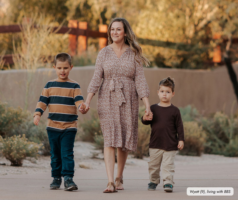 Frame-9469 A woman is walking outdoors with two young boys, each holding one of her hands. She is wearing a patterned dress, while the boy on the left is in a striped long-sleeve shirt and the boy on the right is in a brown long-sleeve shirt and beige pants. A small label at the bottom right of the image reads: “Wyatt (9), living with BBS.”