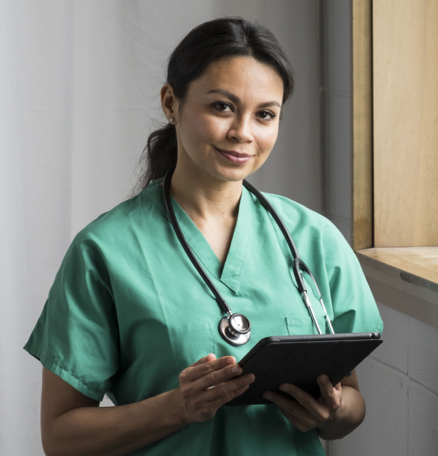 Toni Allen Nurse Image 1 A person wearing green scrubs and a stethoscope around their neck is holding a tablet. They are standing indoors, near a window. A small label at the bottom right states "Actor portrayal.”