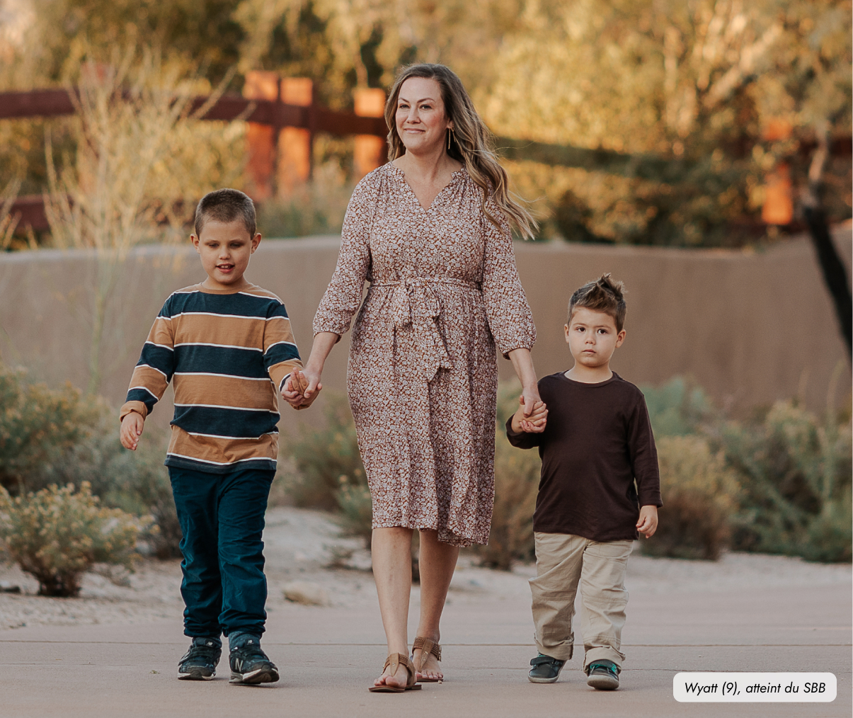 Frame 9457 Une femme marche en extérieur avec deux jeunes garçons qui tiennent chacun une de ses mains. Elle porte une robe à motifs tandis que le garçon à gauche est en tee-shirt à manches longues à rayures et le garçon à droite en tee-shirt à manche longue marron et pantalon beige. Une petite étiquette en bas à droite de l’image dit : « Wyatt (9), atteint du SBB. »